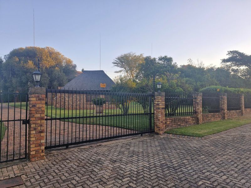 Front view of a residential property featuring a secure metal gate, brick perimeter wall, paved driveway, and a charming thatched roof building.