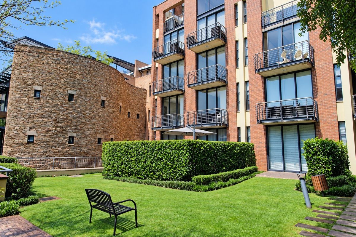 A well-maintained apartment complex exterior featuring brick architecture, private balconies, and a landscaped communal garden area.