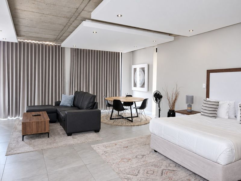 Modern open-plan studio apartment featuring an exposed concrete ceiling, recessed lighting, tiled floors, and floor-to-ceiling curtains.