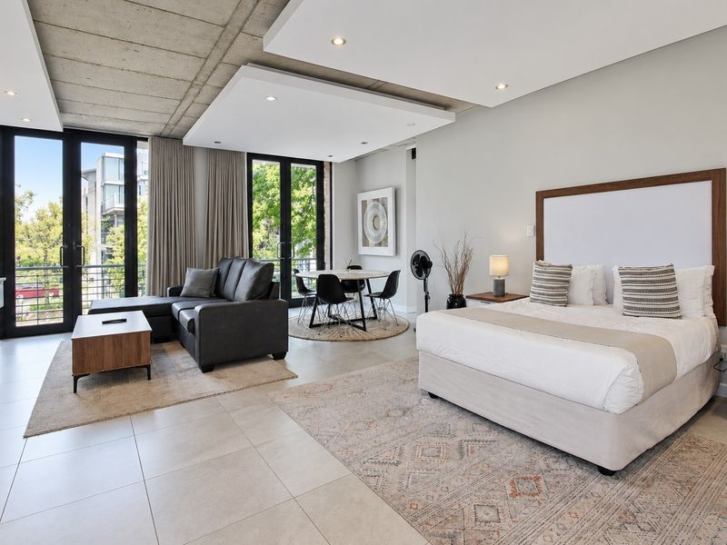 Modern open-plan studio apartment featuring exposed concrete ceilings, tiled floors, and large glass doors leading to a balcony, creating a bright, contemporary living space.