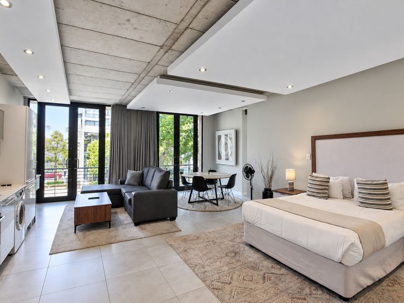 Modern open-plan studio apartment featuring an exposed concrete ceiling, sleek kitchen cabinetry with integrated appliances, and floor-to-ceiling glass doors leading to a balcony.