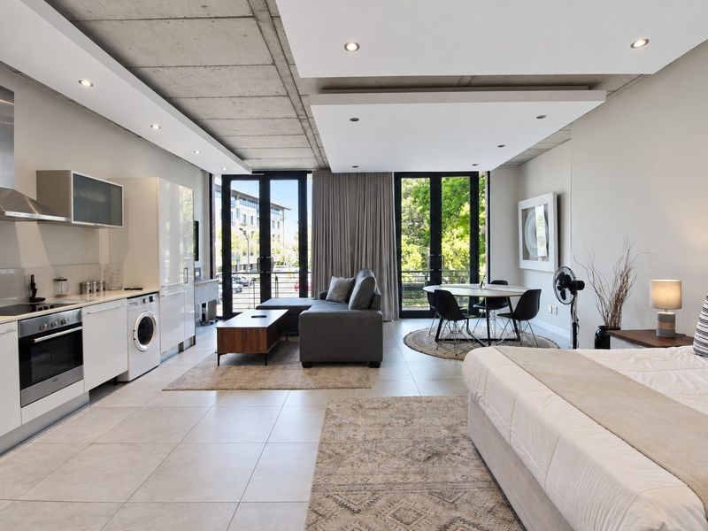 Modern open-plan studio apartment featuring a sleek kitchen with integrated appliances, exposed concrete ceilings, and direct access to a balcony through glass doors.