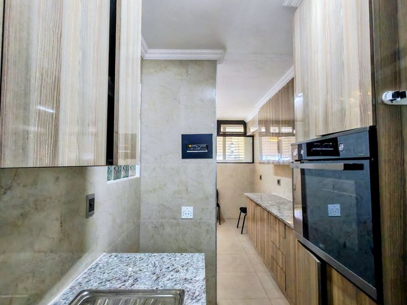 A modern galley-style kitchen featuring wood-grain cabinetry, granite countertops, a built-in oven, and neutral tiled finishes throughout.