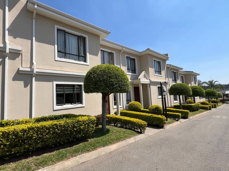 A well-maintained townhouse exterior featuring a beige facade, neat landscaping, paved driveway, and modern aluminium window frames under a clear blue sky.
