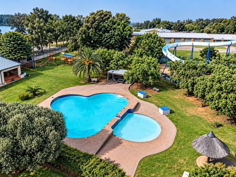 Aerial view of a recreational pool area featuring a large swimming pool, water slide, landscaped gardens, and a thatched gazebo.