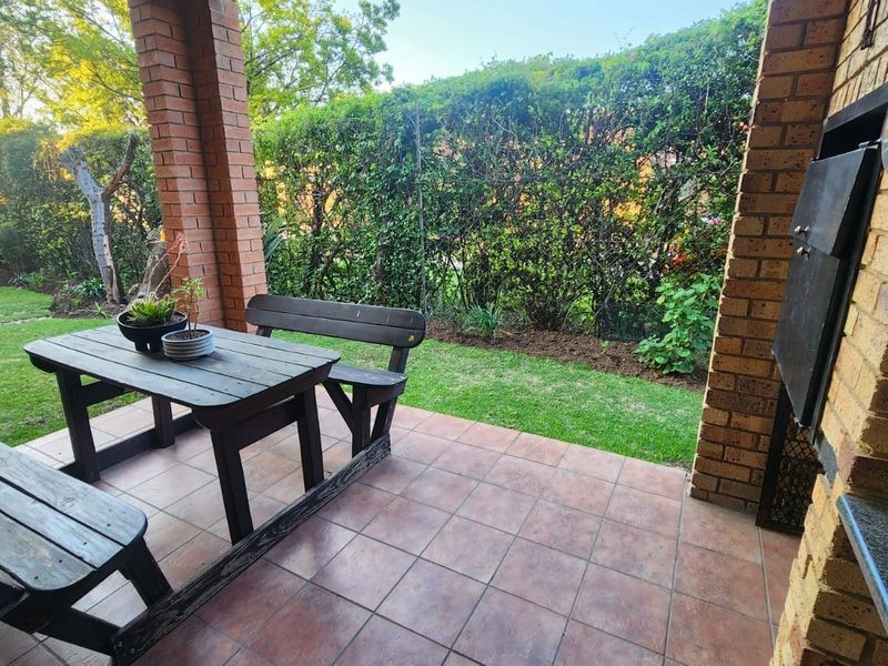 A covered patio area featuring a built-in brick braai, tiled flooring, and brick pillars, overlooking a private garden with established hedging.