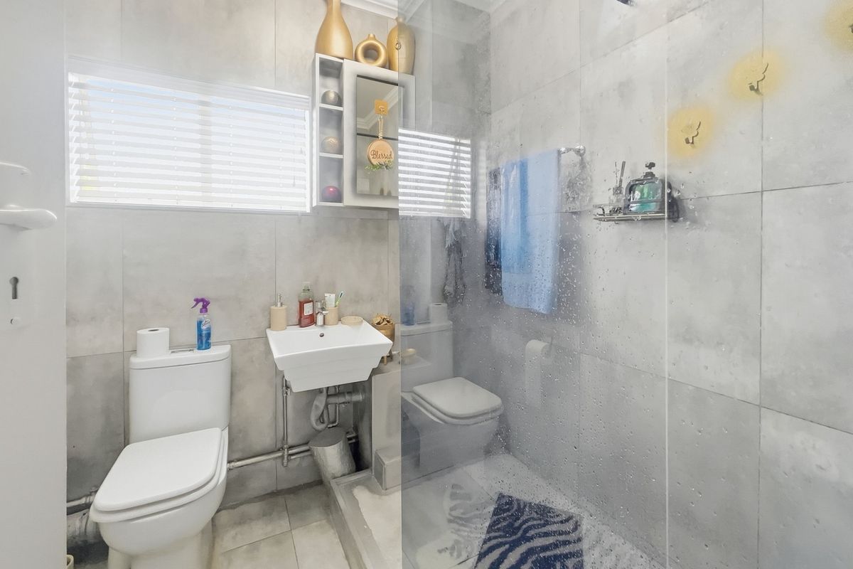 A compact modern bathroom featuring grey wall tiles, a wall-mounted basin, a toilet, and a glass-enclosed shower area.