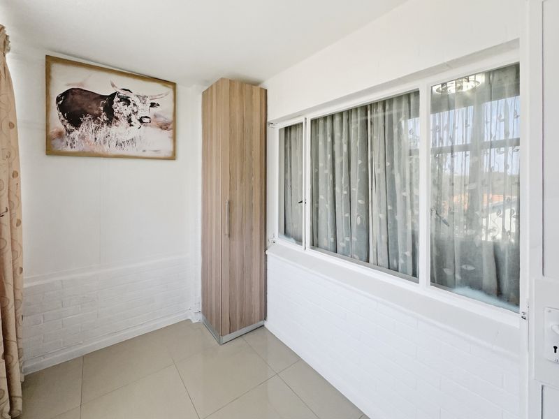 A bright, empty bedroom featuring tiled flooring, painted brick walls, and a built-in wooden cupboard with large windows providing natural light.