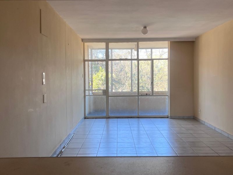 An empty, sunlit living area featuring tiled flooring and a large glass window wall providing natural light and outdoor views.