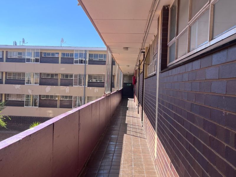 An exterior apartment access walkway featuring tiled flooring, brick walls, and a metal balustrade overlooking the building complex.