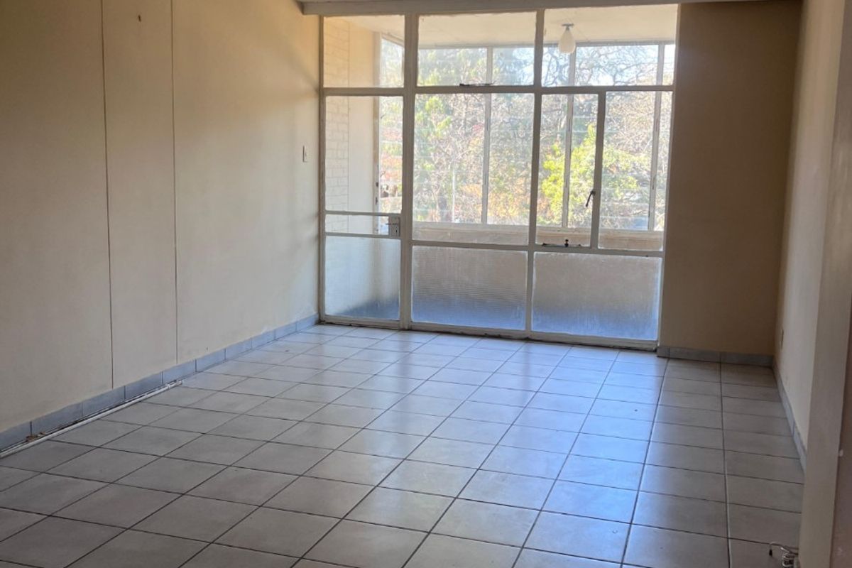An empty, sunlit living area featuring neutral tiled flooring and a large glass partition leading to an enclosed balcony.