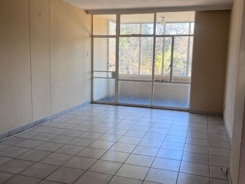 An empty, sunlit living area featuring neutral tiled flooring and a large glass partition leading to an enclosed balcony.