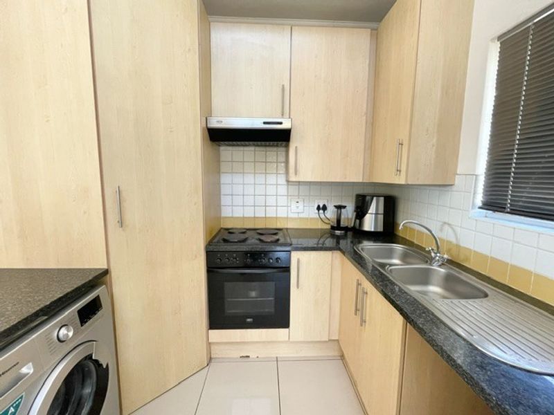 Compact kitchen featuring light wood-finish cabinetry, dark laminate countertops, a built-in oven and hob, and a stainless steel double sink with tiled backsplash.
