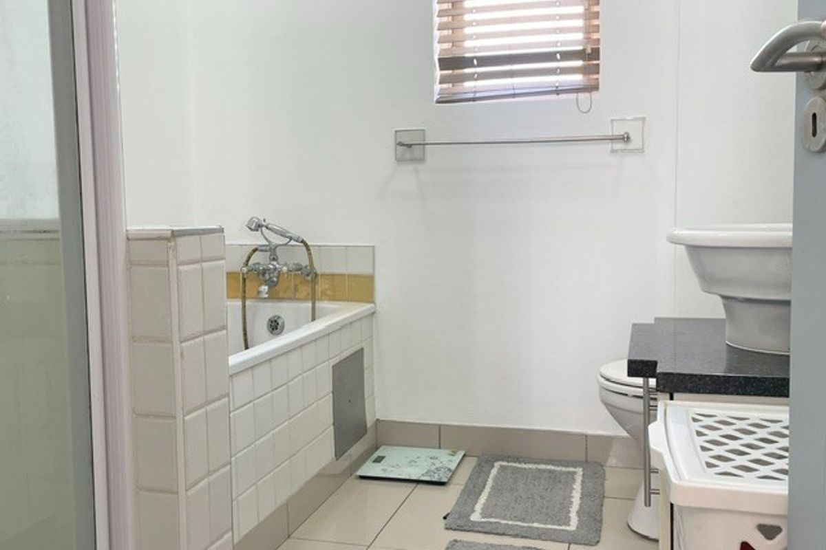 A functional bathroom featuring a bathtub with tiled surround, pedestal basin, and toilet, finished with neutral floor tiles and a window blind.