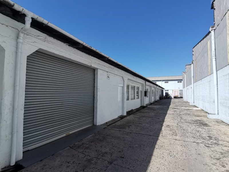Exterior view of an industrial warehouse unit featuring a roller shutter door, concrete driveway, and functional guttering system.