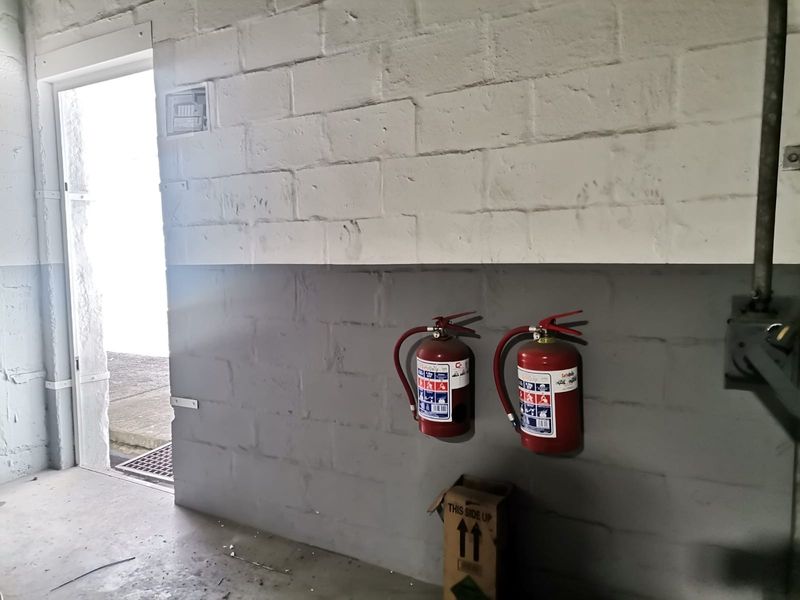 Utility area featuring painted concrete block walls, mounted fire extinguishers, and an electrical distribution board, providing functional industrial storage space.
