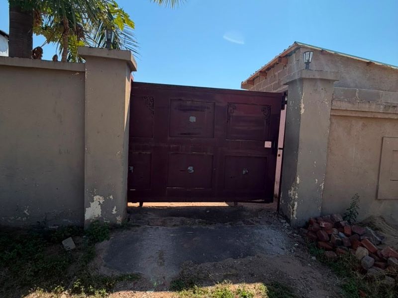Front entrance featuring a solid driveway gate, perimeter walling, and concrete pillars, showing signs of wear and unfinished exterior brickwork.