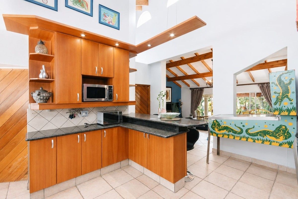 Open-plan kitchen featuring warm wooden cabinetry, granite countertops, a breakfast bar, and exposed timber ceiling beams, flowing into the adjacent living area.