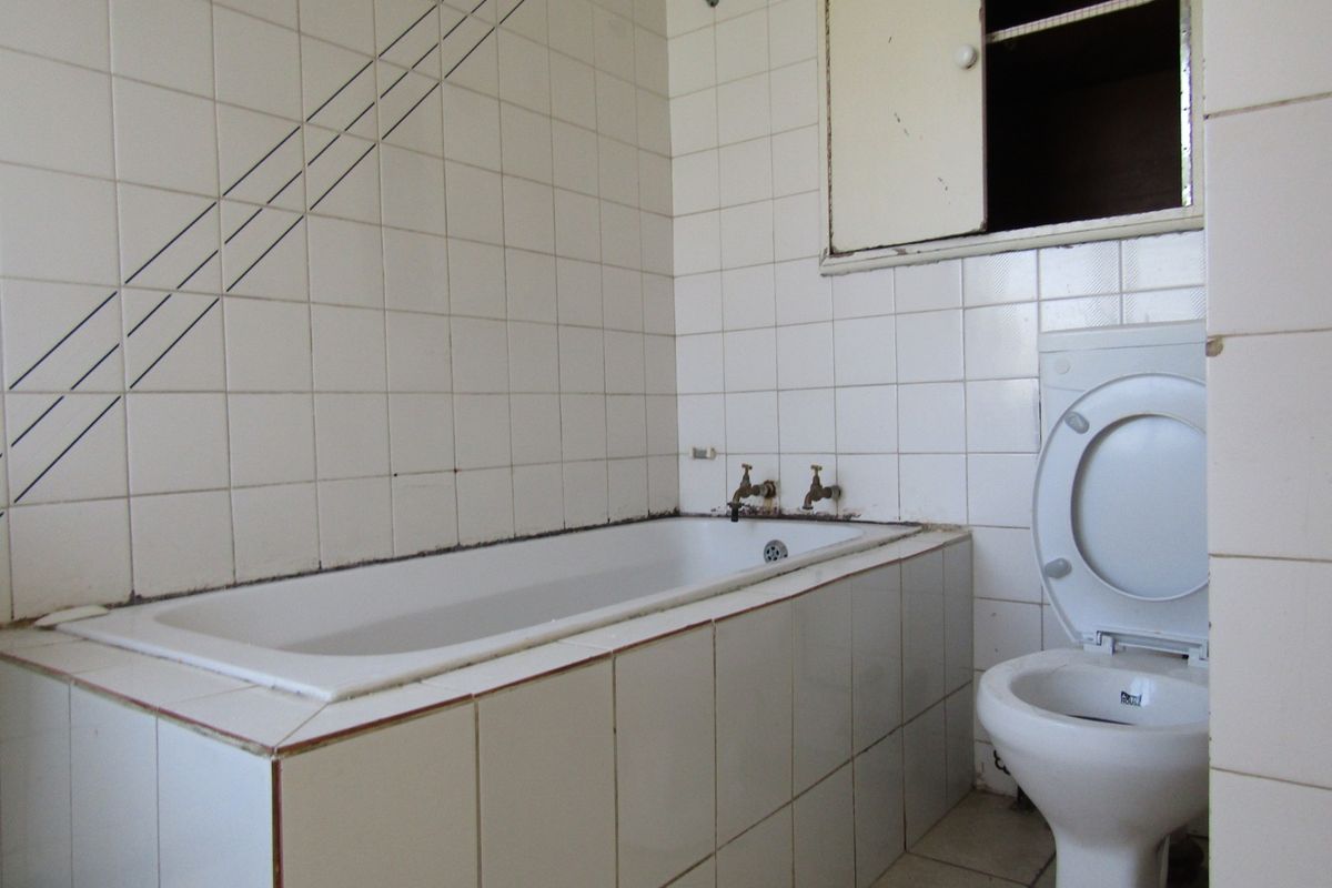 A dated bathroom featuring a built-in bathtub, wall-mounted toilet, and tiled finishes, requiring renovation to update the fixtures and overall condition.