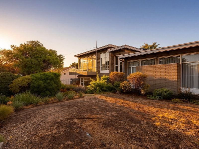Rear view of a modern brick house featuring large windows, an elevated balcony, and a landscaped garden at sunset.