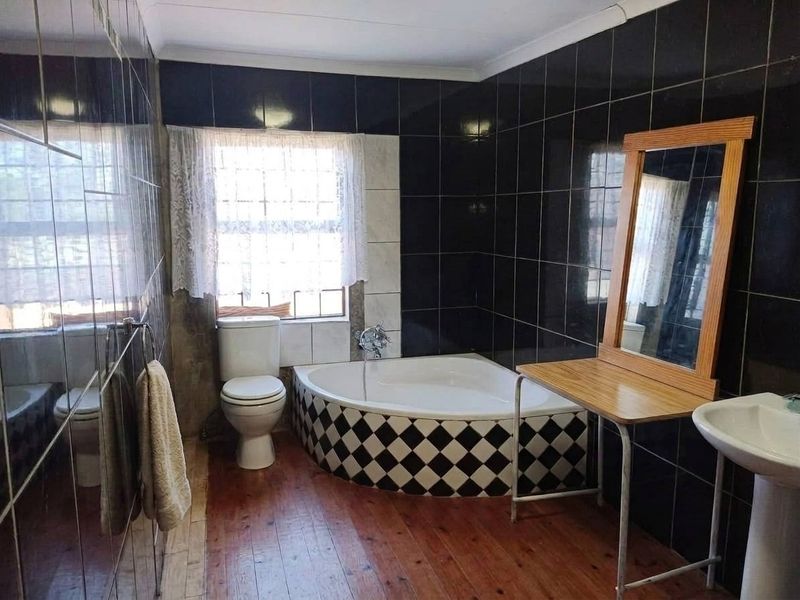 A dated bathroom featuring a corner bathtub, pedestal basin, toilet, and dark wall tiling with timber flooring.