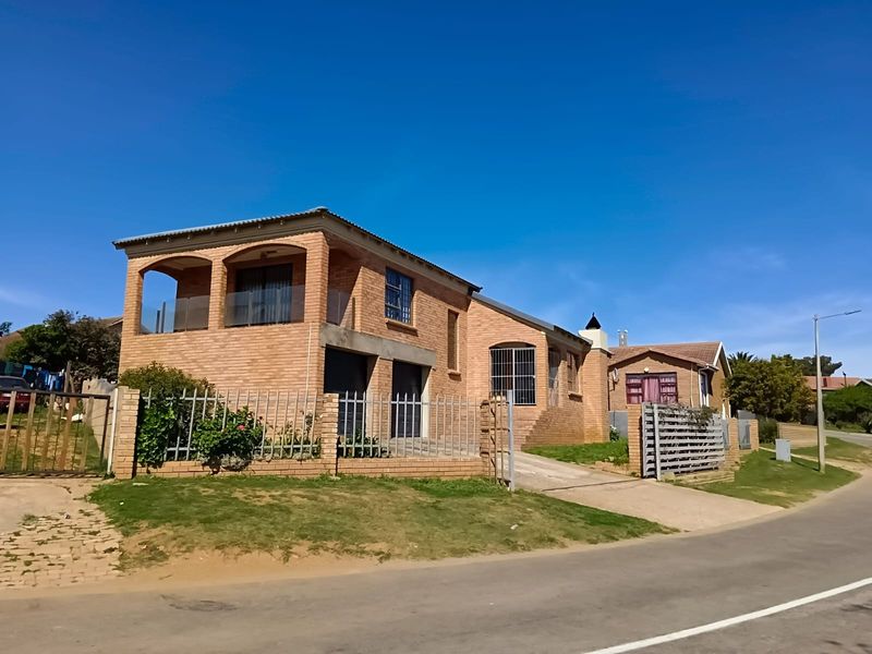A street-facing view of a face brick house featuring a covered balcony with glass balustrades, a paved driveway, and secure perimeter fencing.