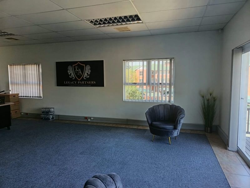 A spacious, open-plan office area featuring carpeted flooring, suspended ceilings with recessed lighting, and large windows with vertical blinds.