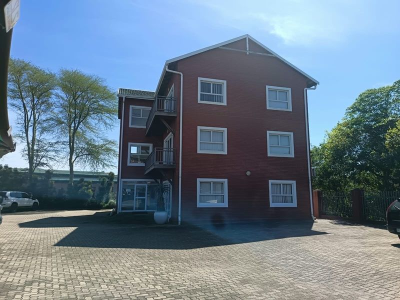 A multi-story office building featuring a red brick facade, multiple balconies, and a spacious paved parking area.
