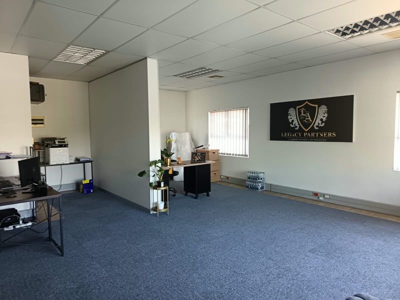 A functional office space featuring carpeted flooring, suspended ceilings with recessed lighting, and large windows with vertical blinds.