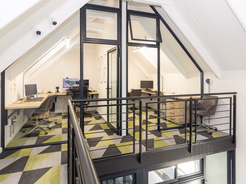 Modern mezzanine office space featuring glass partitions, built-in desks, and contemporary carpet tiles under a vaulted ceiling.