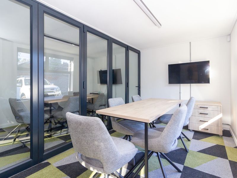 A modern office meeting room featuring glass partitions, patterned carpet tiles, and wall-mounted display screen.