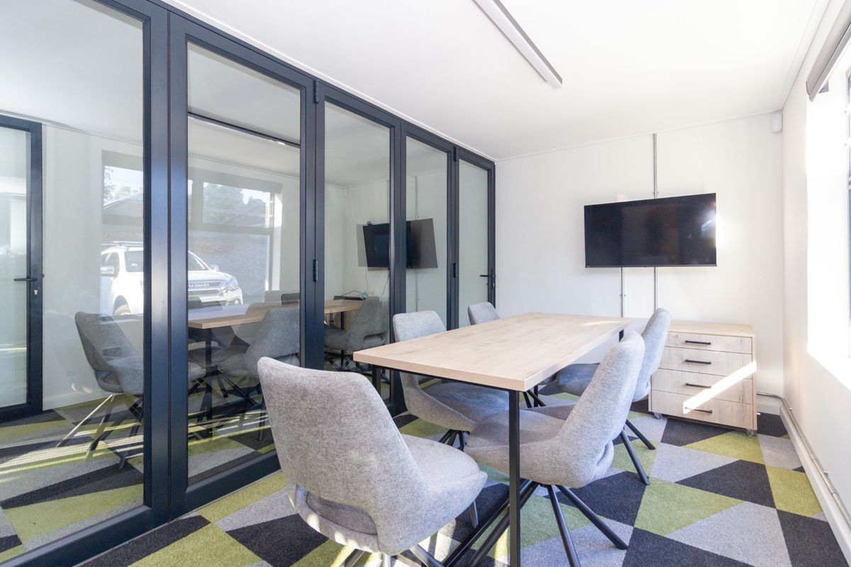 A modern office meeting room featuring glass partitions, patterned carpet tiles, and wall-mounted display screen.