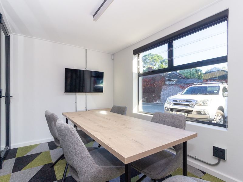 A modern, well-lit office space featuring patterned carpet tiles, large aluminium-framed windows, and surface-mounted electrical infrastructure.