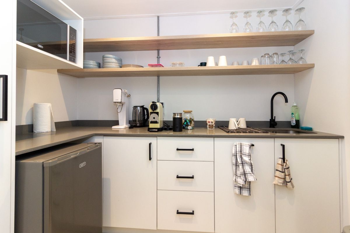 A modern, compact kitchenette featuring white cabinetry, stone countertops, floating timber shelving, and a sleek matte black faucet.