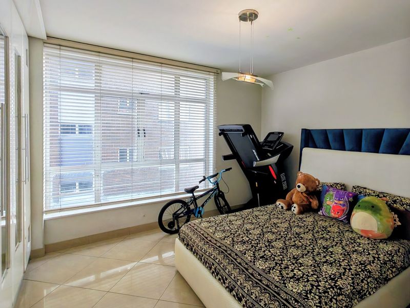 A well-lit bedroom featuring tiled flooring, built-in cupboards, and large windows with venetian blinds, offering a clean and functional living space.