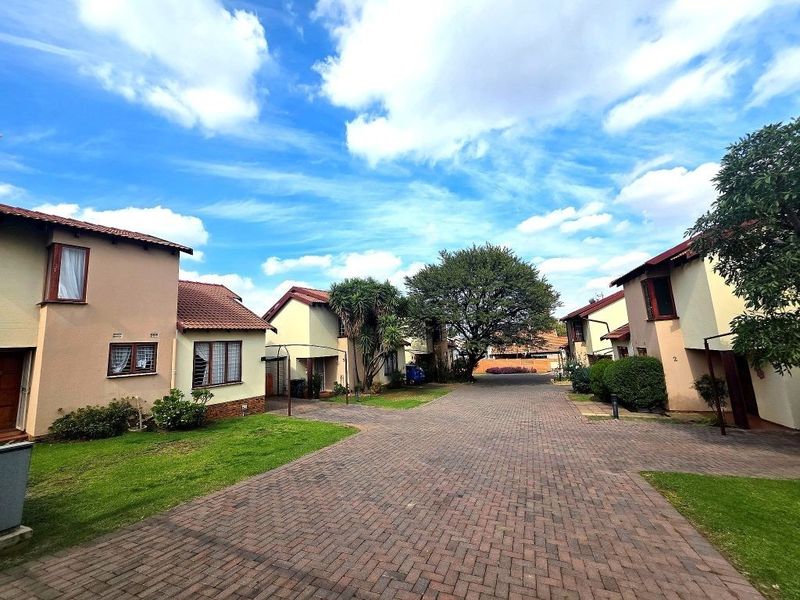 A view of the townhouse complex entrance featuring a paved driveway, landscaped lawns, and multiple residential units with tiled roofs.