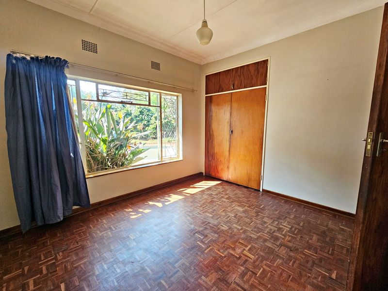 A bright, empty bedroom featuring original parquet flooring, a built-in wooden wardrobe, and large windows providing natural light and garden views.
