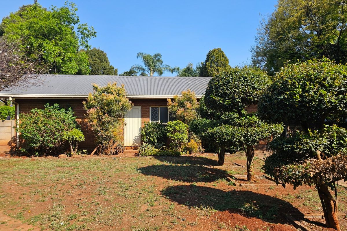 A view of the rear brick facade of the house, featuring a corrugated metal roof and a well-established garden with mature trees.