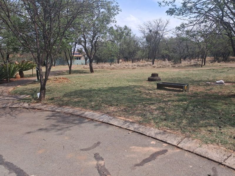 A view of a vacant residential plot featuring a grass lawn, mature trees, and a paved curb along the street edge.