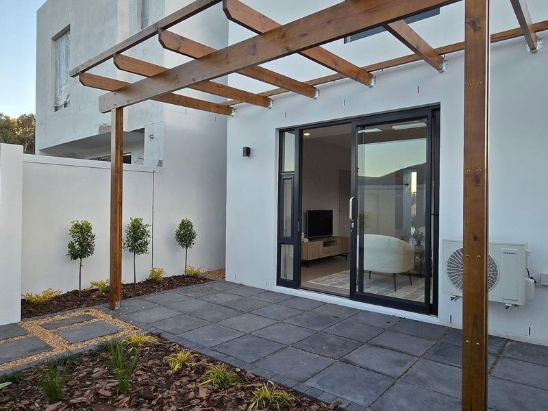 Modern outdoor patio featuring a wooden pergola, paved flooring, built-in braai area, and direct access to the living space through sliding glass doors.