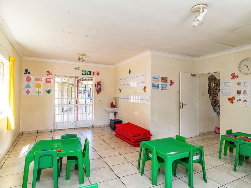 A functional interior space currently utilized as a classroom, featuring tiled flooring, a wall-mounted basin, and a secure metal entrance gate.