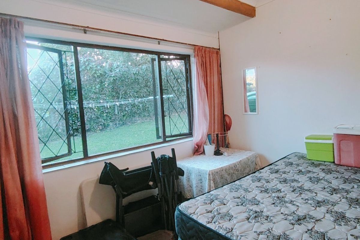 A bedroom featuring tiled flooring, an exposed timber ceiling beam, and a large window with diamond-patterned security bars overlooking the garden.