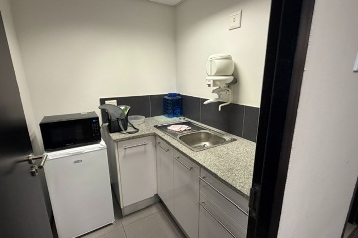 A compact office kitchenette featuring granite countertops, stainless steel sink, built-in cabinetry, and a tiled backsplash.
