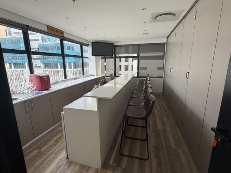 A modern, well-maintained office bar area featuring sleek built-in cabinetry, stone countertops, and large windows providing ample natural light and city views.