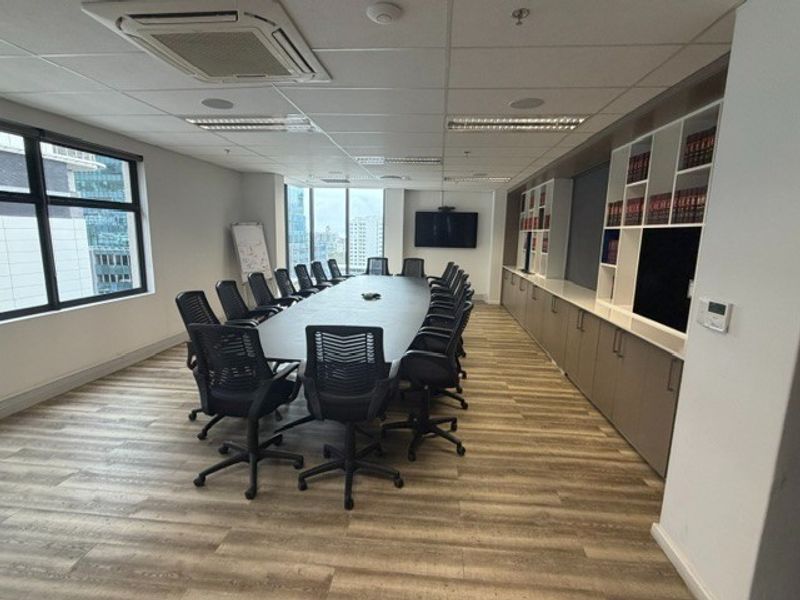 A professional boardroom featuring a large conference table, built-in storage cabinetry, suspended ceiling with integrated lighting, and large windows with city views.