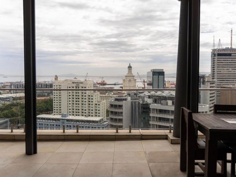 A spacious covered balcony featuring tiled flooring and a modern glass balustrade, offering expansive elevated city and harbour views.