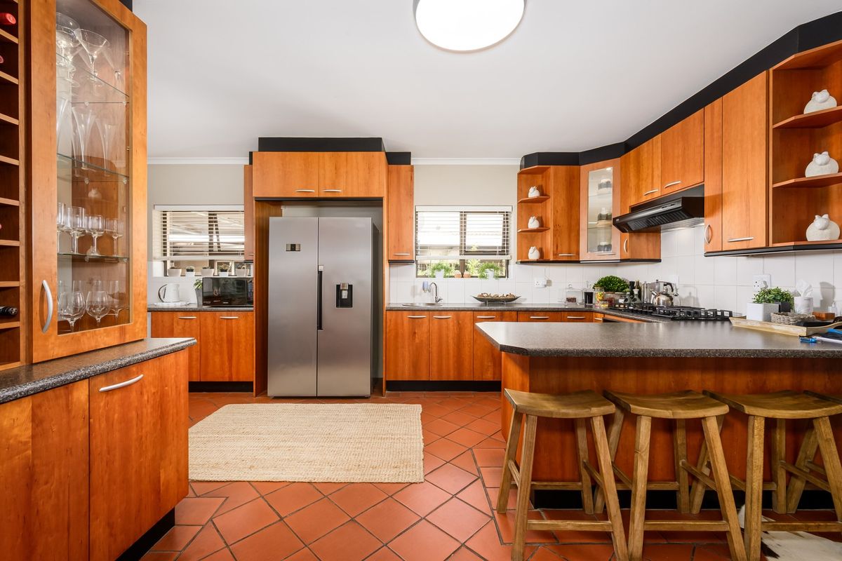 A functional kitchen featuring warm-toned wooden cabinetry, dark laminate countertops, a gas hob with extractor, and durable terracotta-style tiled flooring.