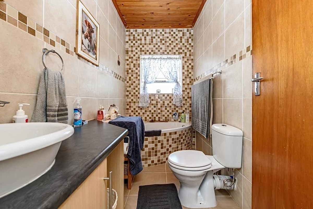 A compact bathroom featuring a built-in vanity with a vessel sink, a tiled bathtub, and a toilet, finished with neutral wall tiles and a timber ceiling.