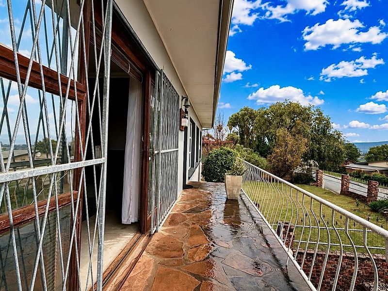 A covered balcony featuring stone paving, metal security gates, and a protective railing, offering an elevated view of the surrounding garden and neighborhood.