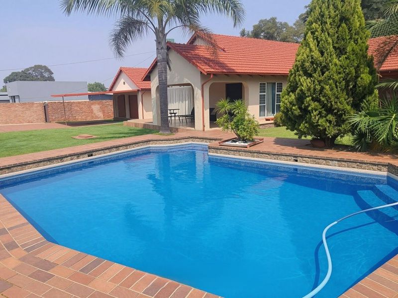 A well-maintained swimming pool with brick coping, set in a landscaped garden with a covered patio and tiled roof house exterior.